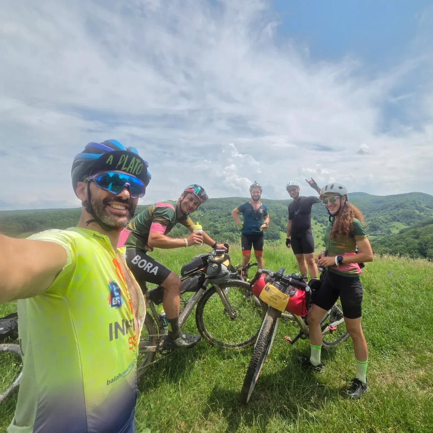 Six bikepackers grinning on a hilltop meadow, loaded bikes in the grass and forested hills behind