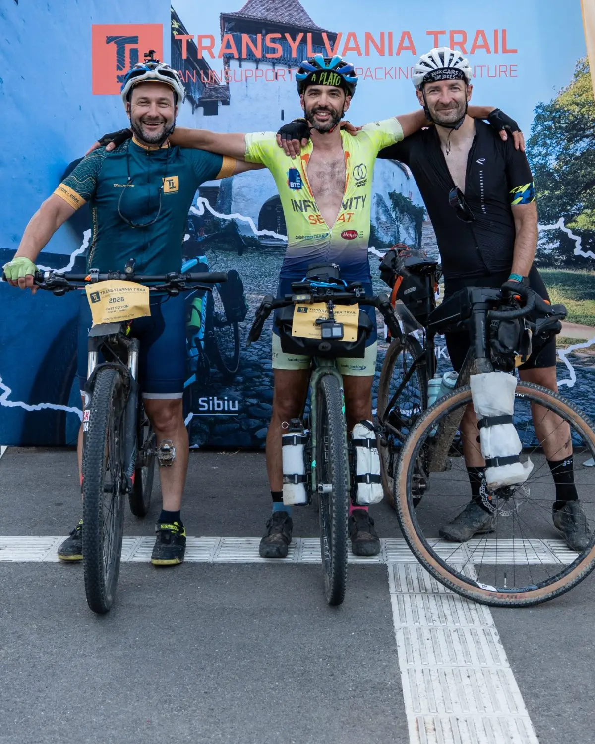 Three riders with muddy bikes, arms around each other at the Transylvania Trail finish in Sibiu.