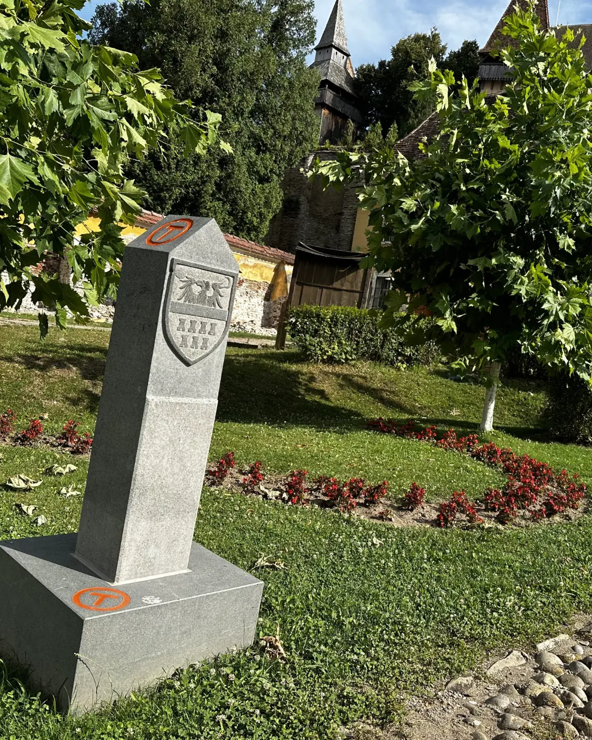 A carved Via Transilvanica milestone with the orange T and a coat of arms, on the green below the fortified church in Biertan.