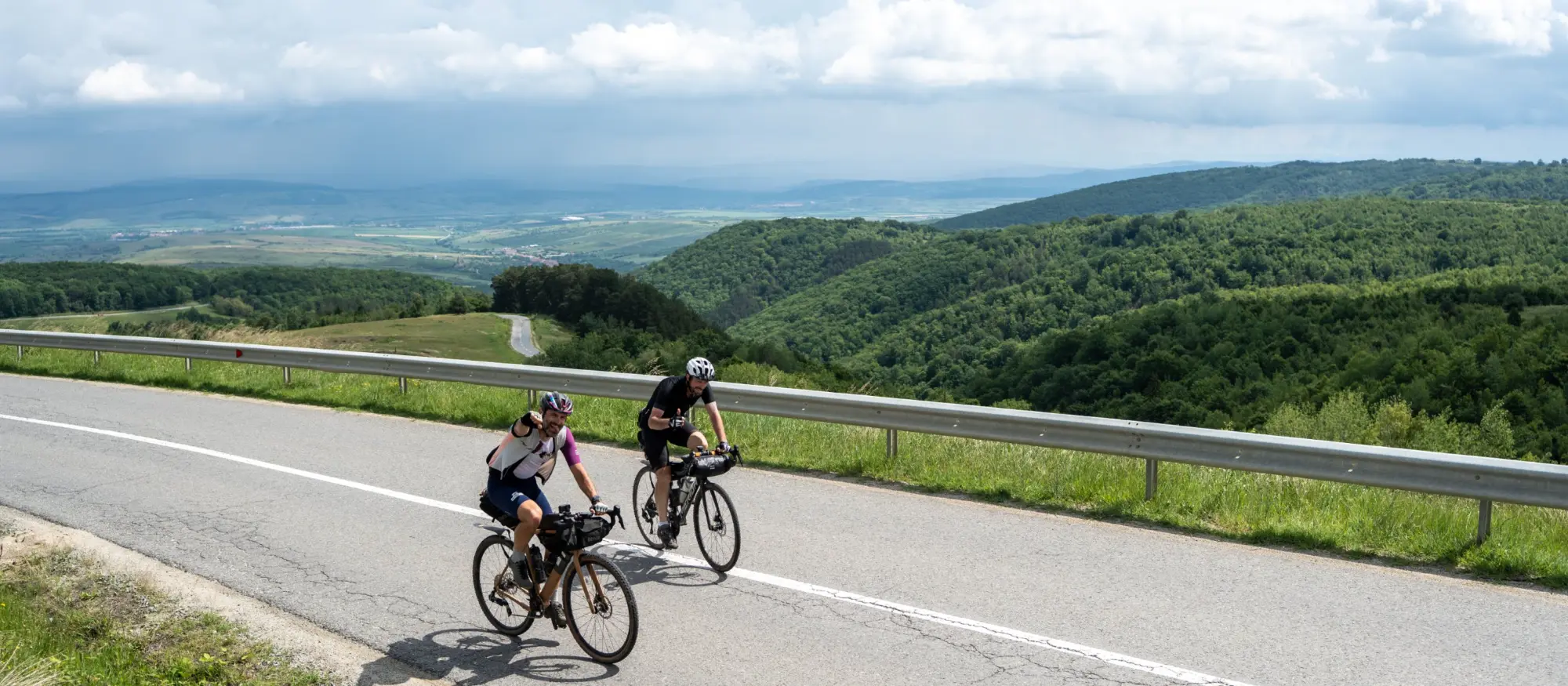 Two riders climbing a tarmac pass road with the forested hills and plain of southern Transylvania opening out behind them.