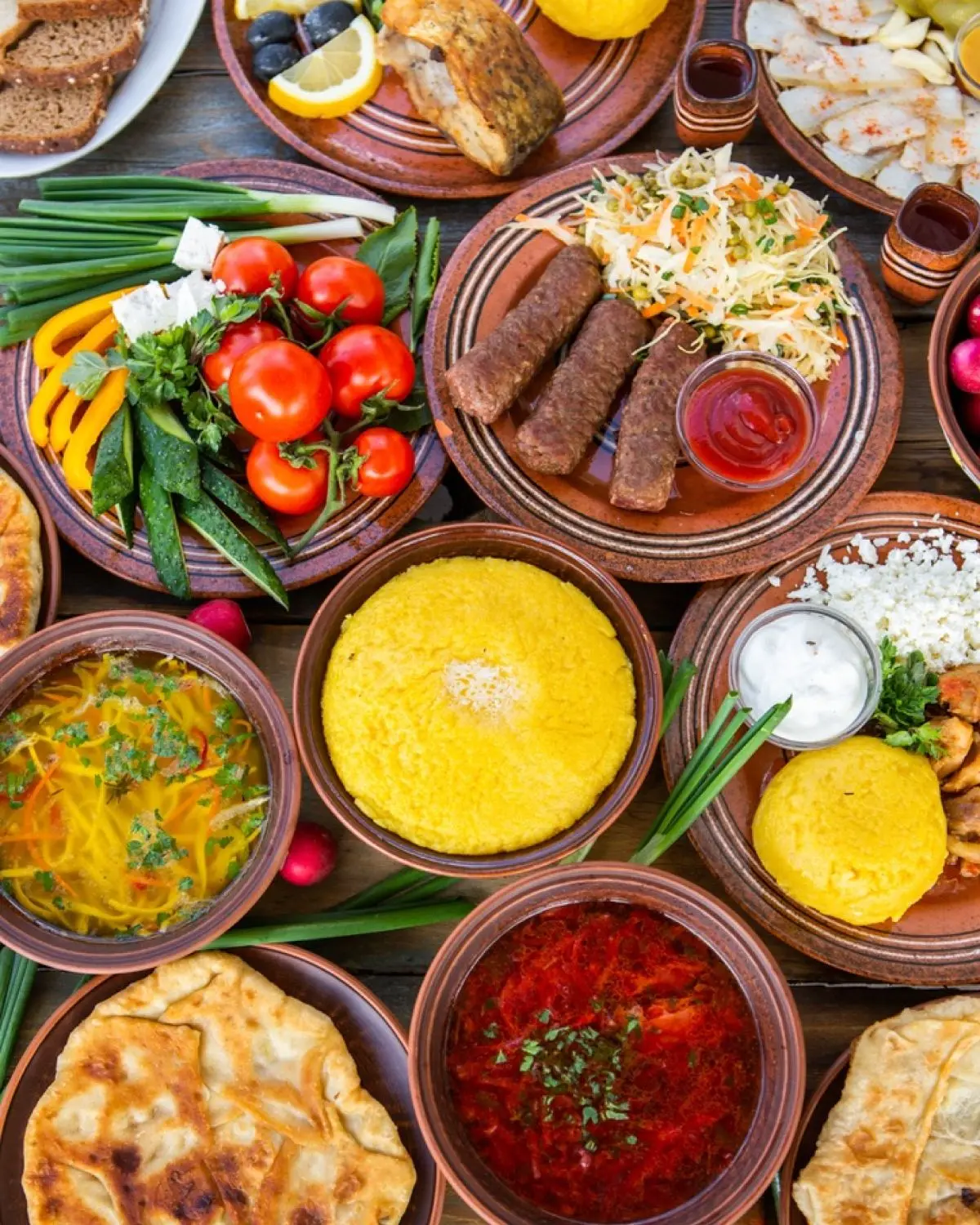 A table of Romanian dishes from above: polenta, ciorbă, mici, cheese and fresh vegetables.